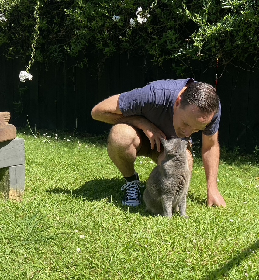 Steve Mullan crouching with a grey cat in a sunny garden